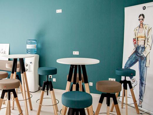 Gebäude 2 In der Au - Moderner Pausenraum mit mehreren Hochtischen und Hockern, sowie einem Aufstellen mit einer Modezeichnung.