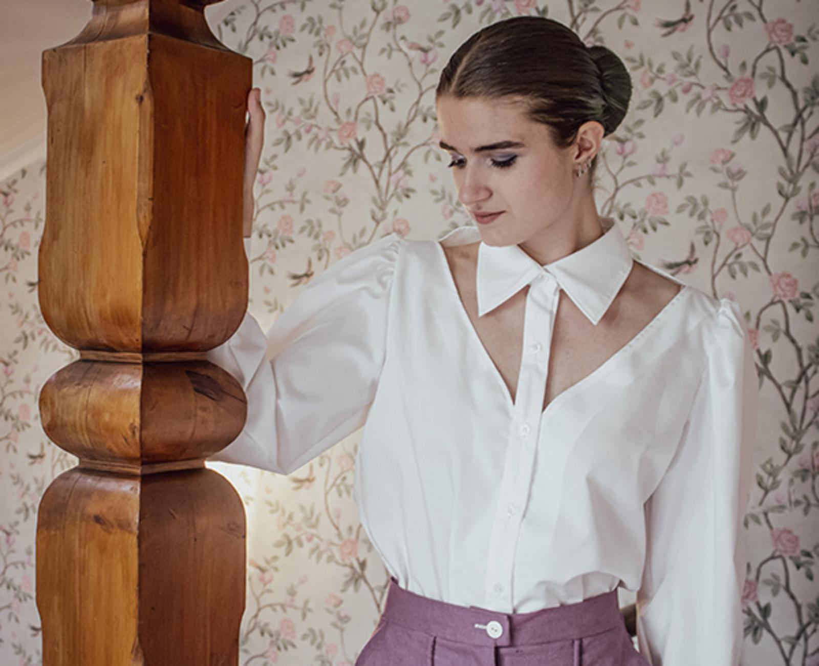 Person in weißer Bluse und lila Hose lehnt an Holzsäule