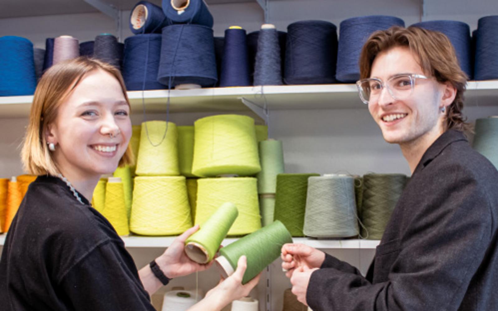 Zwei Personen halten Garnrollen vor einem Regal mit verschiedenfarbigen Garnkegeln