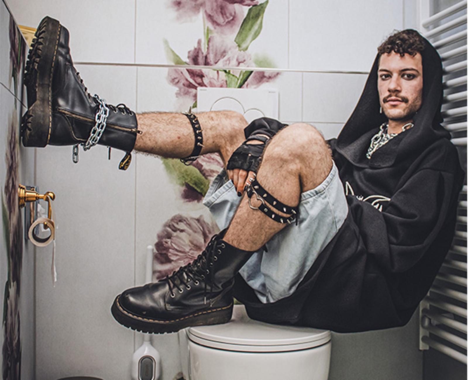 Person im schwarzen Hoodie und Boots posiert sitzend auf Toilettendeckel